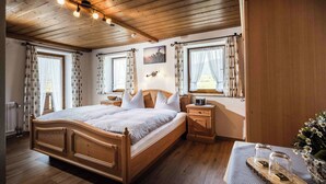 Een bed met houten frame, een witte spreidek en kussens, een nachtkastje met een radio en een raam met gordijnen.