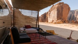 Una autocaravana con una zona de estar exterior cubierta, un sofá, una mesita y vistas a un paisaje desértico con formaciones rocosas.