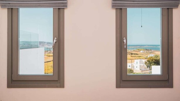 Dues finestres amb persianes grises, cadascuna amb vista a un paisatge costaner i edificis.