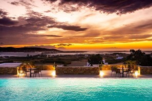 A poolside area with tables and chairs, overlooking a scenic landscape with a sunset. - Kouros Blanc Paros (Paros)