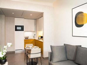 A modern hotel room with a grey sofa, a dining area with a table and chairs, and a kitchen in the background. - Adagio Apart Hotel (Abidjan)