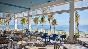 Un espace extérieur aménagé avec des tables et des chaises, offrant une vue sur une plage bordée de palmiers et sur l’océan.