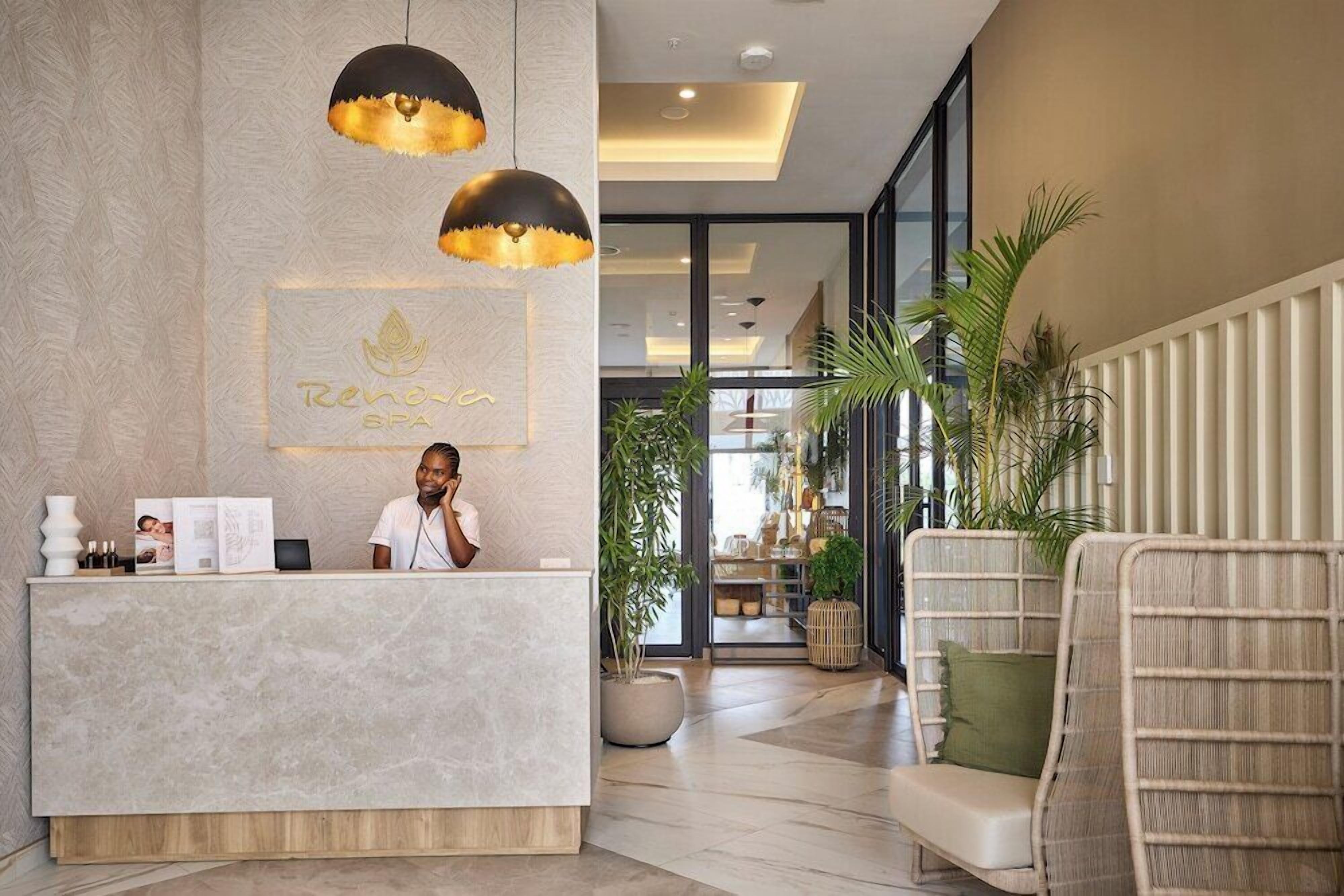 A reception area with a marble counter, a receptionist, and a reception desk. There are hanging pendant lights, a framed sign above the counter, and a plant to the right.