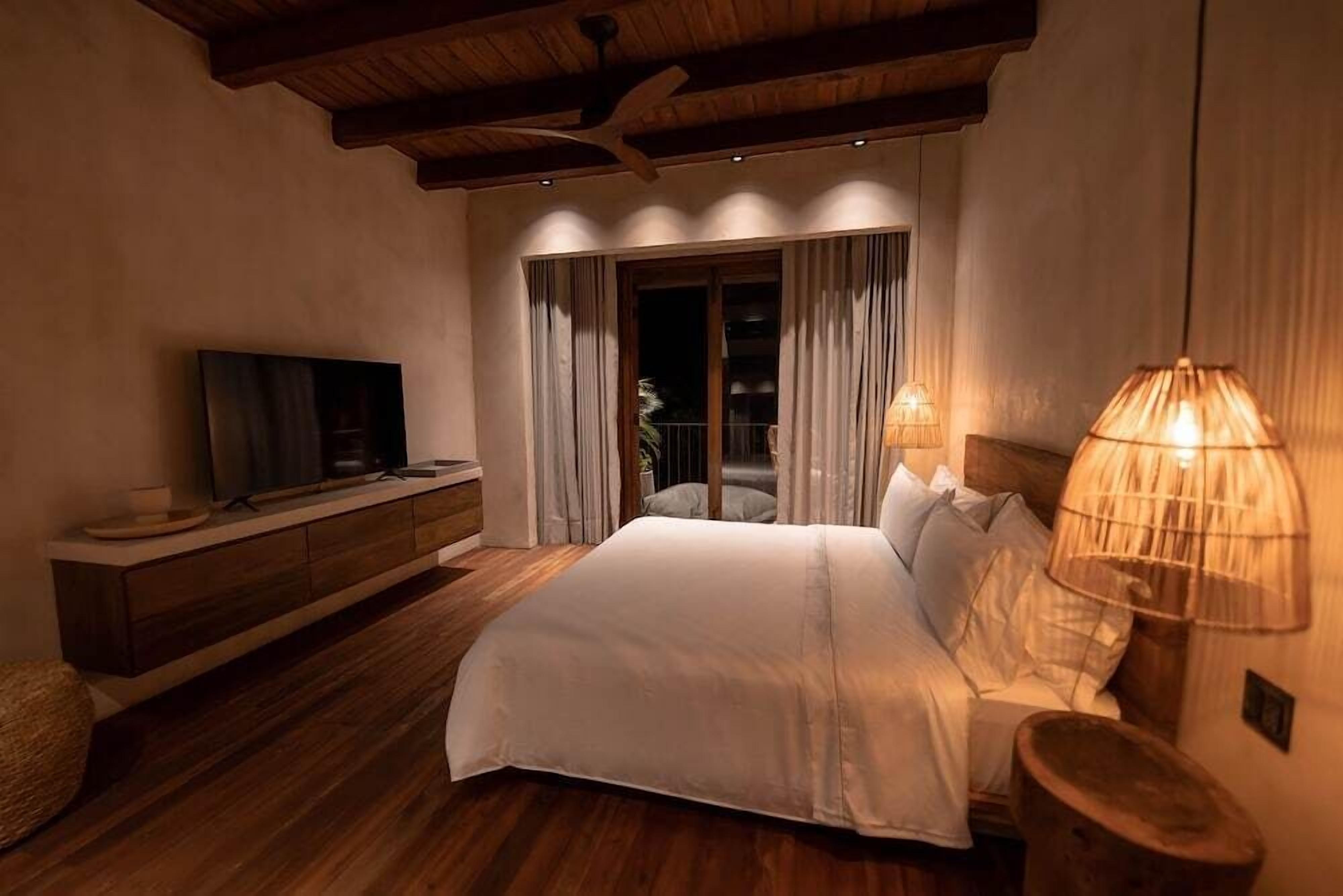 A bedroom with a large bed, wooden flooring, a TV on a wooden console, and a balcony with curtains.