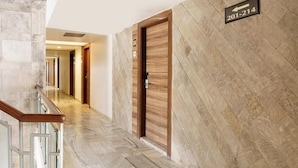 Un couloir d’hôtel moderne avec un sol à motifs en chevrons, une porte en bois portant un numéro et un garde-corps en verre.