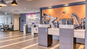 A hotel reception area with a check-in counter, a digital kiosk, and a mural on the wall. - Premier Inn York City Centre (York)