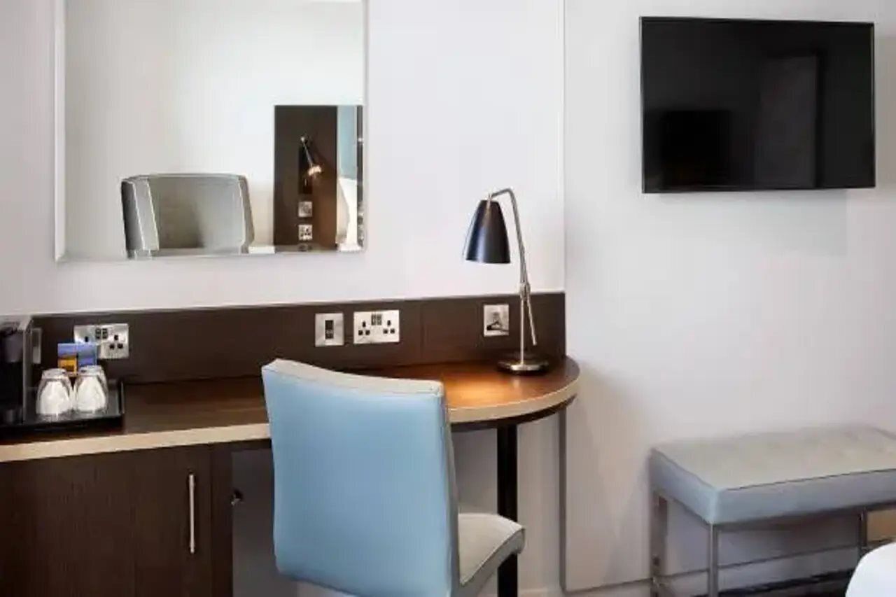 Habitación de hotel con escritorio, silla, mesita de noche, lámpara y televisor de pantalla plana montado en la pared.