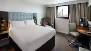 A hotel room with a large bed, a desk with a chair, a chair with a cushion, a lamp, and a window with curtains. - Premier Inn York City Centre (York)