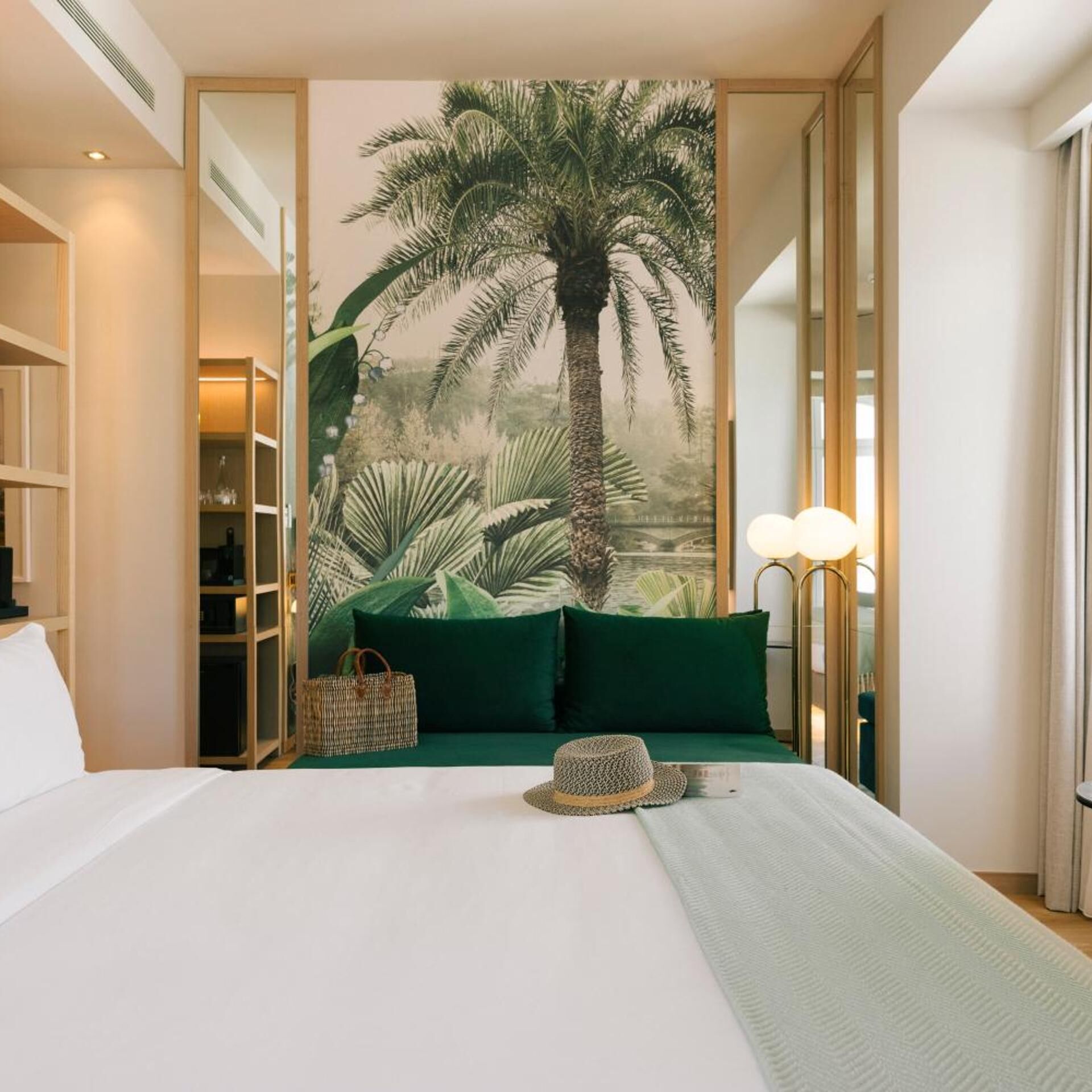 A hotel room with a bed, a large tropical mural, a green sofa, a wicker basket, and a hat.