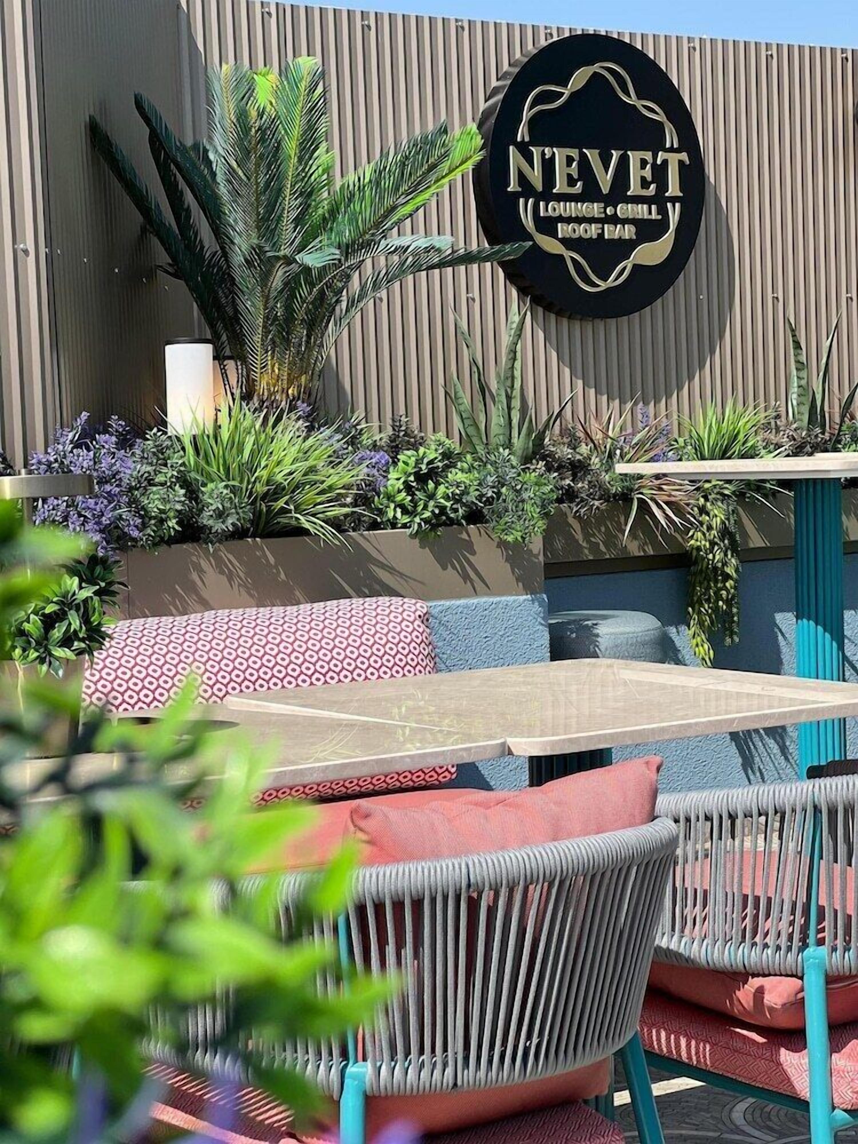 An outdoor seating area with wicker chairs and cushions, a sign for 'NEVET Lounge Grill Rooftop Bar', and a variety of plants and greenery.