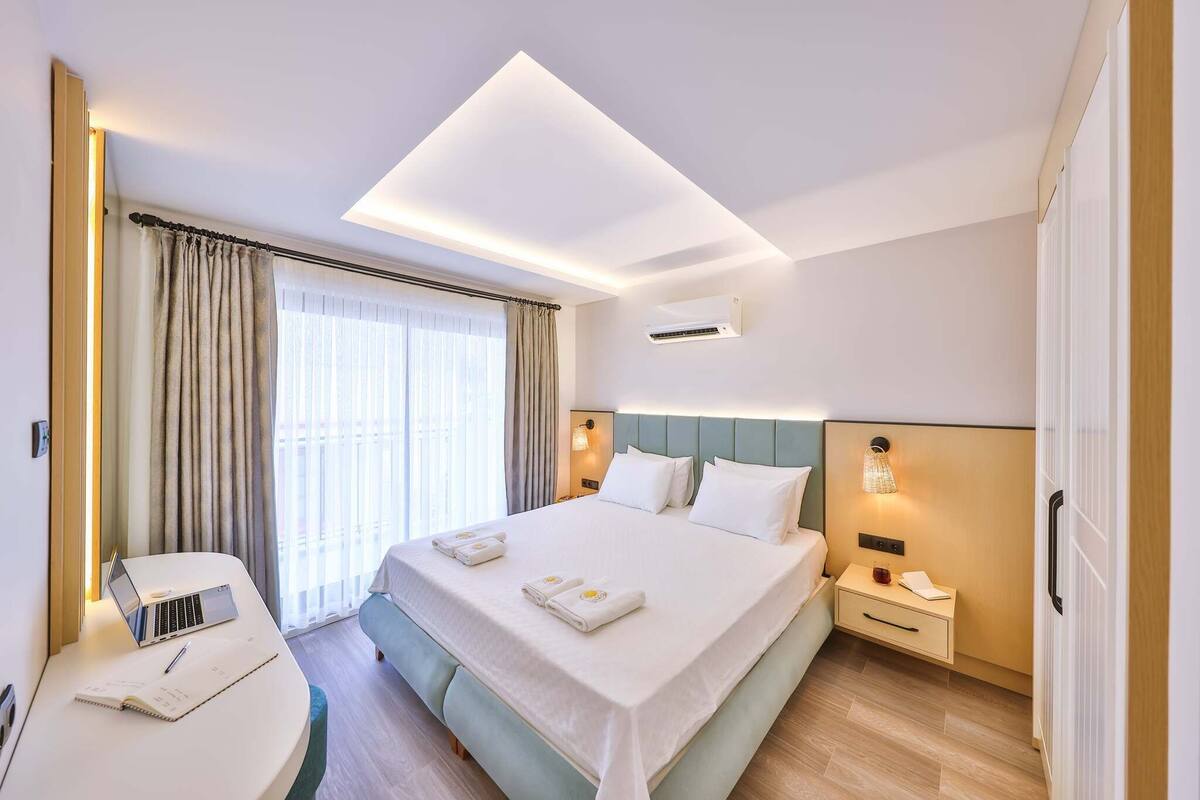 Moderni hotellihuone, jossa on suuri sänky, työpöytä kannettavalla tietokoneella, yöpöytä ja ikkuna, jossa on verhot.