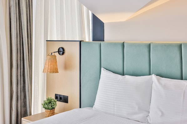 Hotellihuone, jossa on sänky, yöpöytä, seinään kiinnitetty lamppu ja ruukussa oleva kasvi.