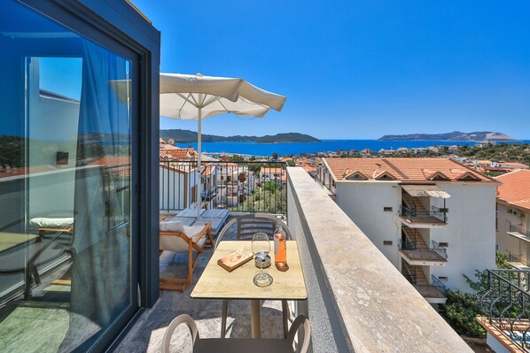 A balcony with a table, chairs, and an umbrella overlooking a coastal town. - Sole Suites (Kaş)