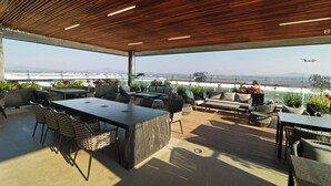 Un patio al aire libre con techo de madera, arreglos de asientos y vista al aeropuerto de fondo.