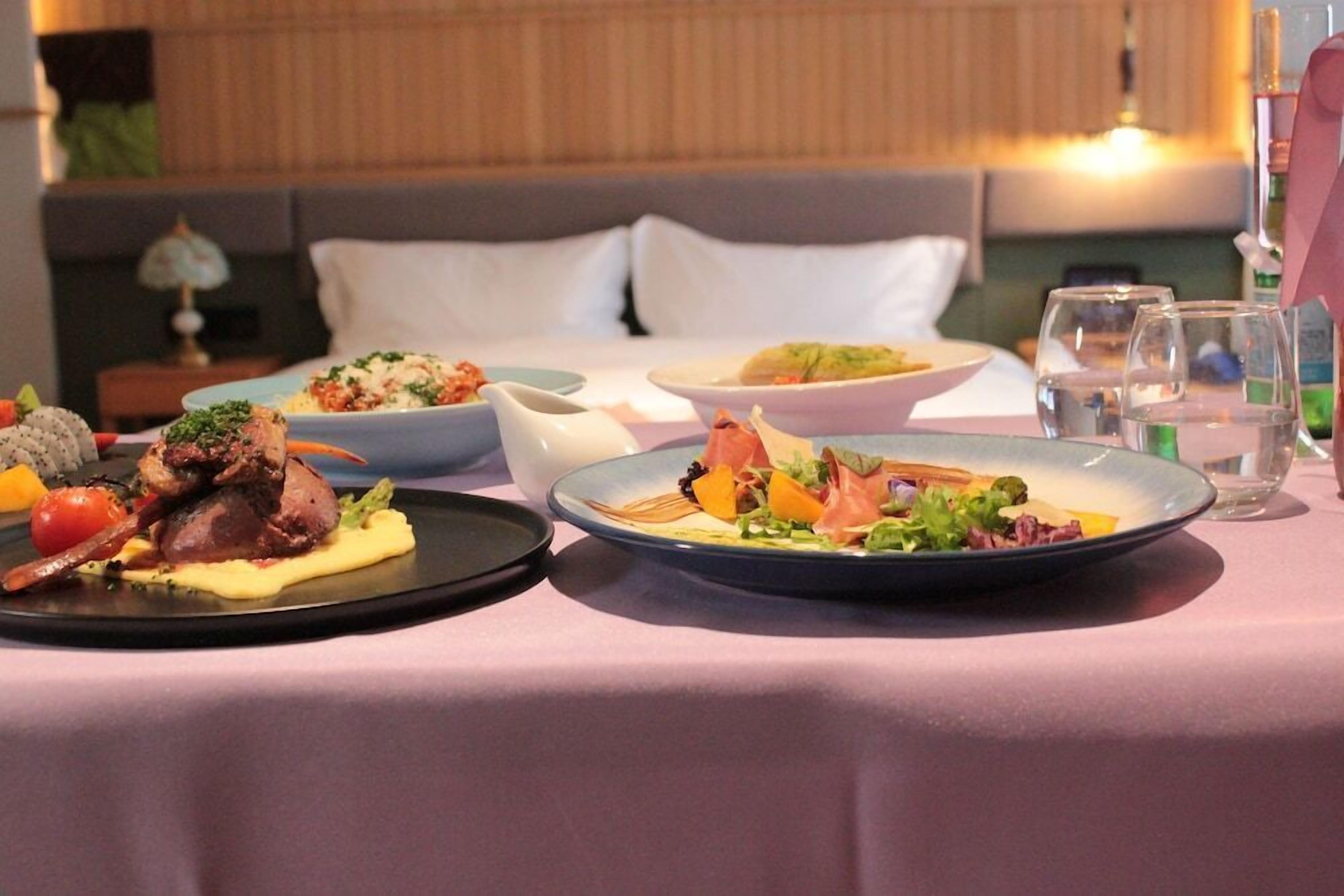 A dining table with plates of food, glasses, and a bed in the background.