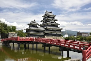 Un castillo japonés tradicional con varios niveles, un puente rojo que lo lleva a él, y una masa de agua al frente.