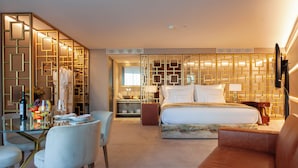 Ein Hotelzimmer mit einem Glastisch, weißen Stühlen, einem Bett mit Kopfteil, einer braunen Couch und einer dekorativen Wandverkleidung.