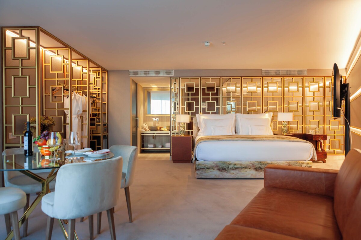 Quarto de hotel com mesa de jantar de vidro, cadeiras brancas, cama com cabeceira, sofá marrom e um painel decorativo na parede.