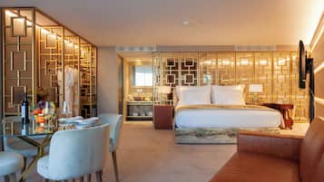 Una habitación de hotel con una mesa de comedor de vidrio, sillas blancas, una cama con cabecera, un sofá marrón y un panel decorativo en la pared.