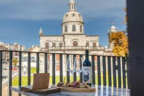 En balkon med et vinflaske, glas og en bog, der kigger ud over en historisk bygning med en kuppel.