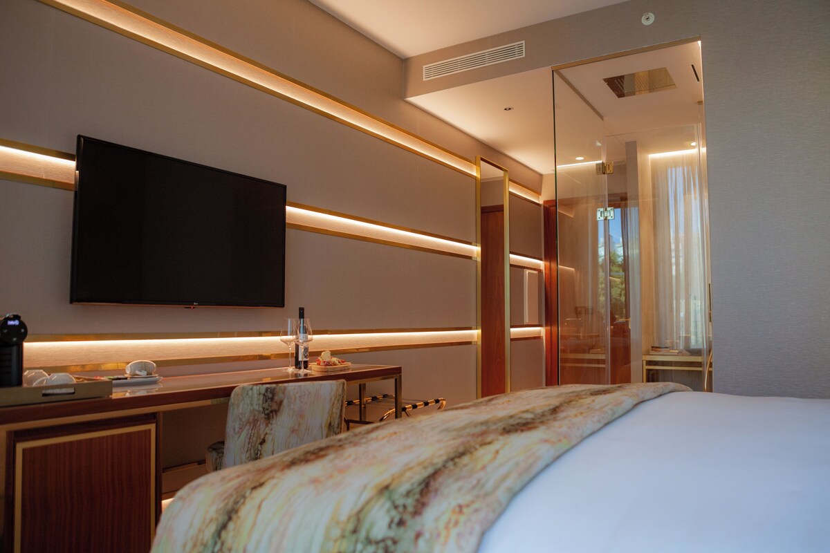 Quarto de hotel com cama, secretária e televisão de tela plana, espelho e chuveiro envidraçado.