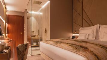 A hotel room with a bed, a desk, a chair, a glass-enclosed shower, and a bedside telephone.