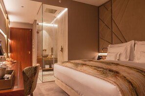 Quarto de hotel com cama, escrivaninha, cadeira, chuveiro em cabine de vidro e telefone ao lado da cama.