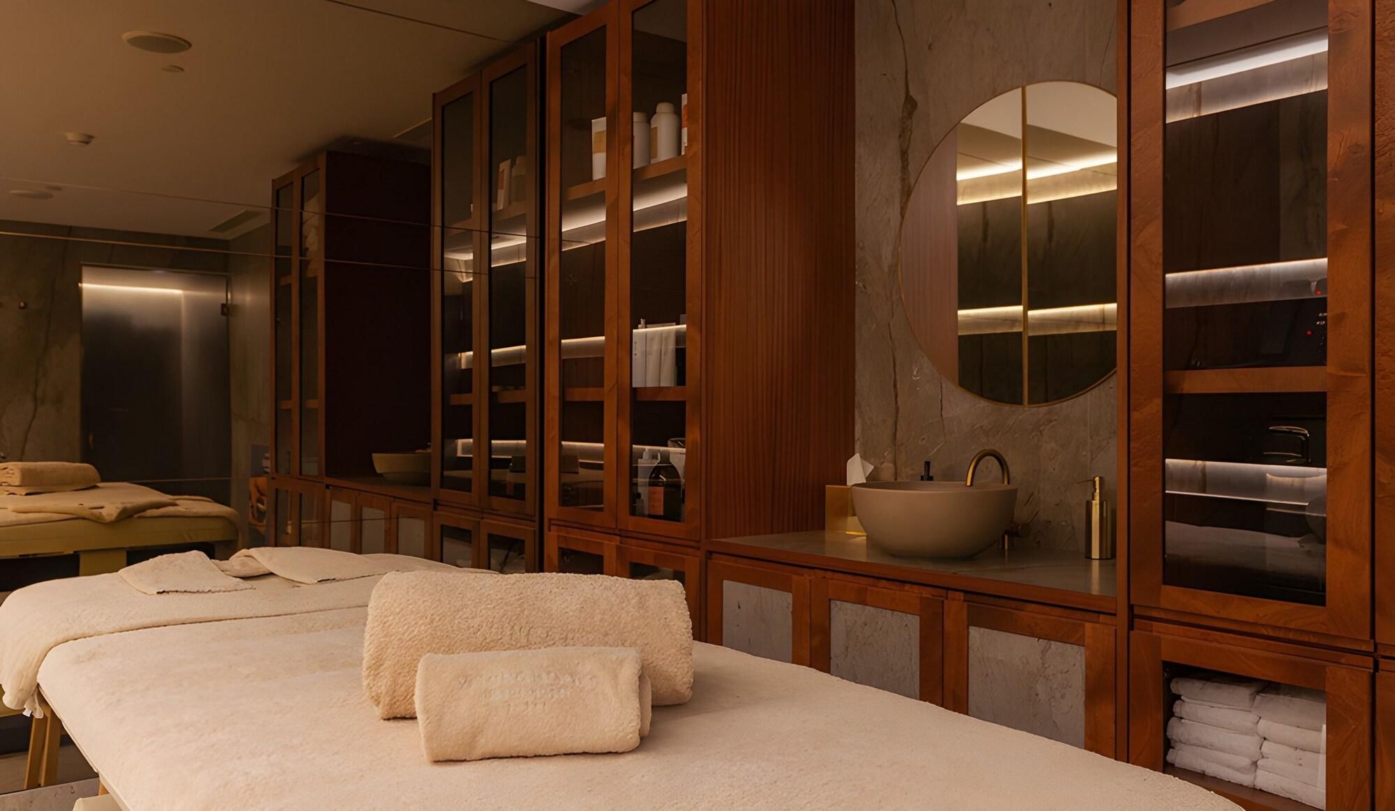Um espaço de spa com mesa de massagem, toalhas, um armário de madeira grande com portas de vidro e um espelho redondo.