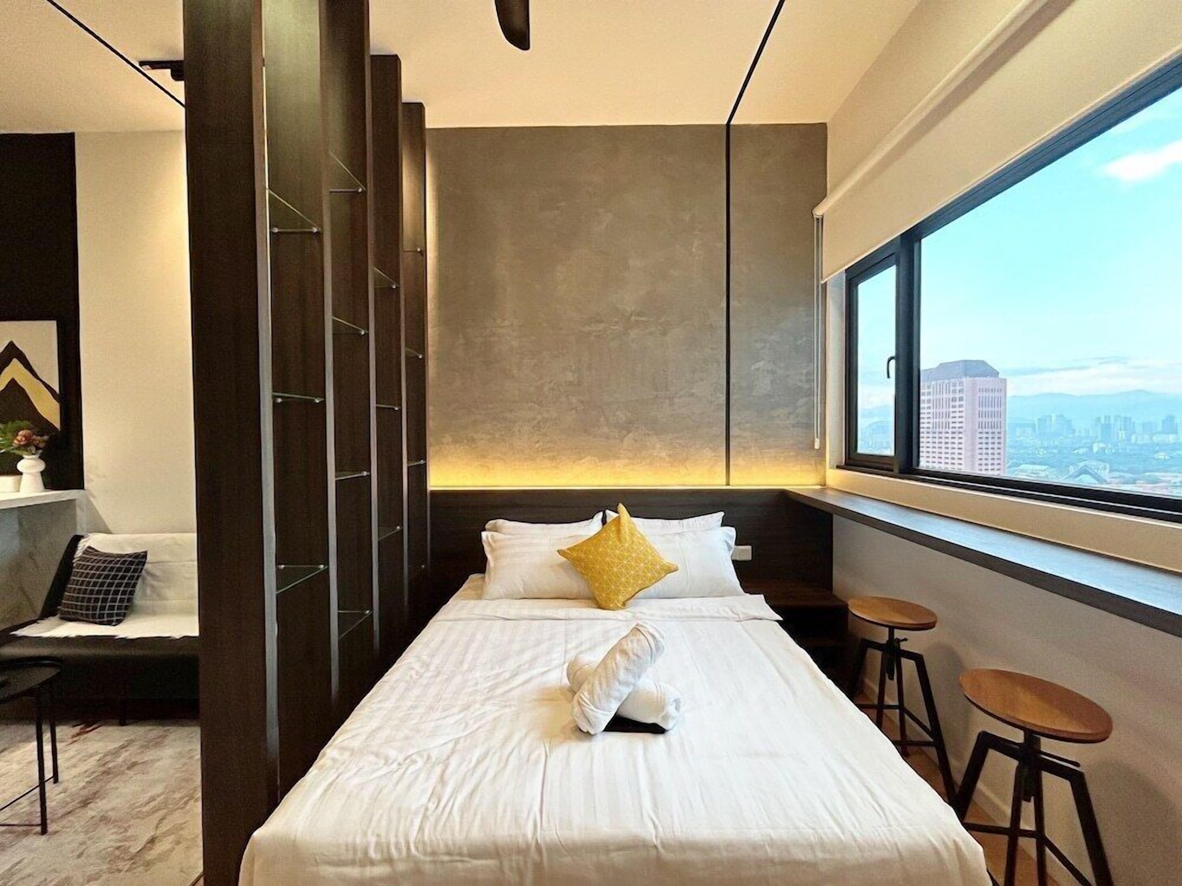 Ein modernes Hotelzimmer mit einem großen Bett, Blick auf die Stadt durch ein großes Fenster, einem eingebauten Kleiderschrank und einer Sitzecke mit Hocker.