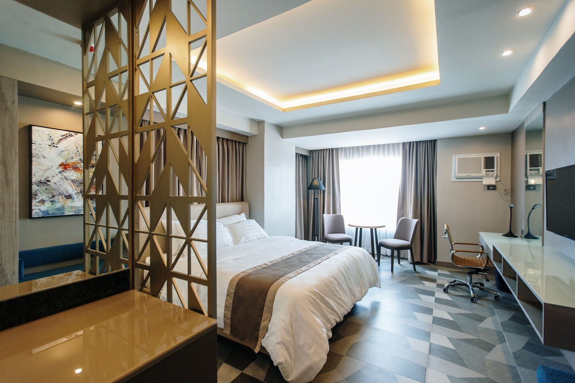A hotel room with a bed, a desk, two chairs, a lamp, and a large window.