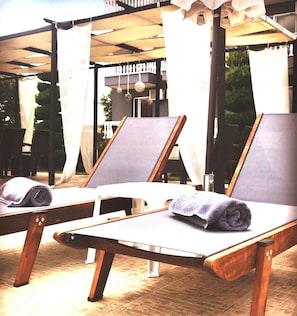 Un espace extérieur ensoleillé, aménagé avec des fauteuils de jardin en bois et une table, doté d’un auvent et offrant une vue sur un bâtiment en arrière-plan.