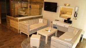 A modern hotel room with a glass-enclosed shower, a flat-screen TV, a minibar, and a seating area with a sofa and ottoman. - Kornilios Palace Thessaloniki (Chalkidona)