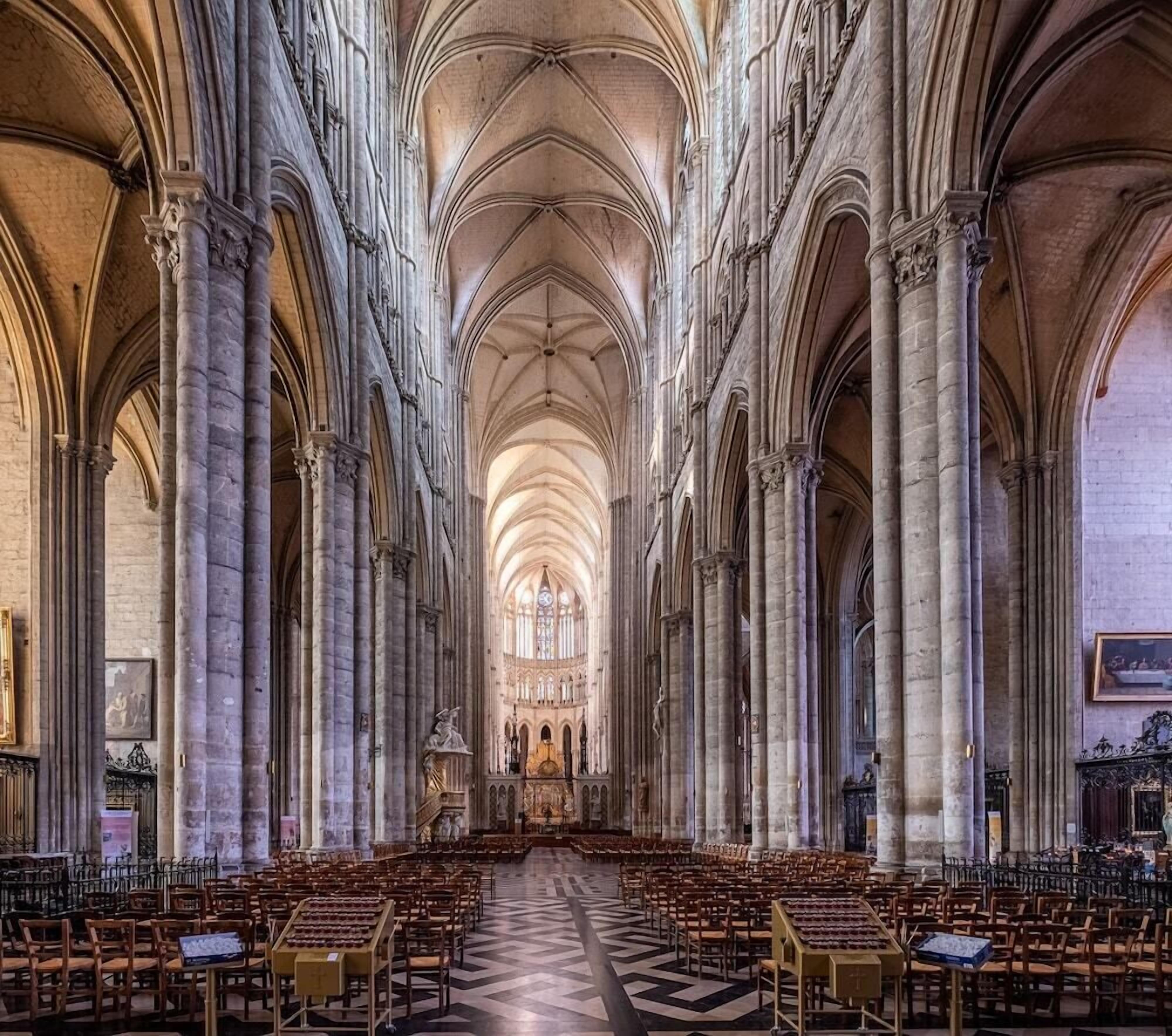 A cathedral interior with high vaulted ceilings, large stone columns, and rows of wooden pews.
