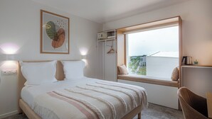 Ein Schlafzimmer mit Bett, Nachttisch, Fenster und Blick auf Gebäude.