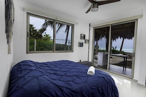 A bedroom with a large window, a bed with a blue cover, a ceiling fan, and a view of a balcony with a table and chairs. - Angelina 28 Beachfront by Nah Hotels (Playa del Carmen)