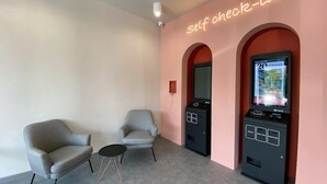 A room with two armchairs, a small round table, a self-check-in kiosk, and a neon sign reading "self check-in". - Woco Hotel Kinrara (Puchong)