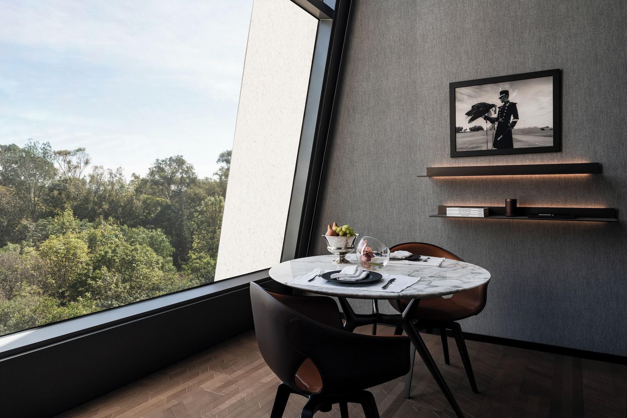 Een moderne eetruimte met een ronde tafel, stoelen, uitzicht op groen buiten, een ingelijst beeld aan de muur en een plank met boeken en een beker.