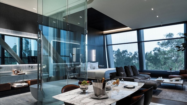 A modern interior with a glass-enclosed bathroom, a dining area with a marble table, and a living space with a sofa and a view of trees. - Hotel Alexander (Mexico City)