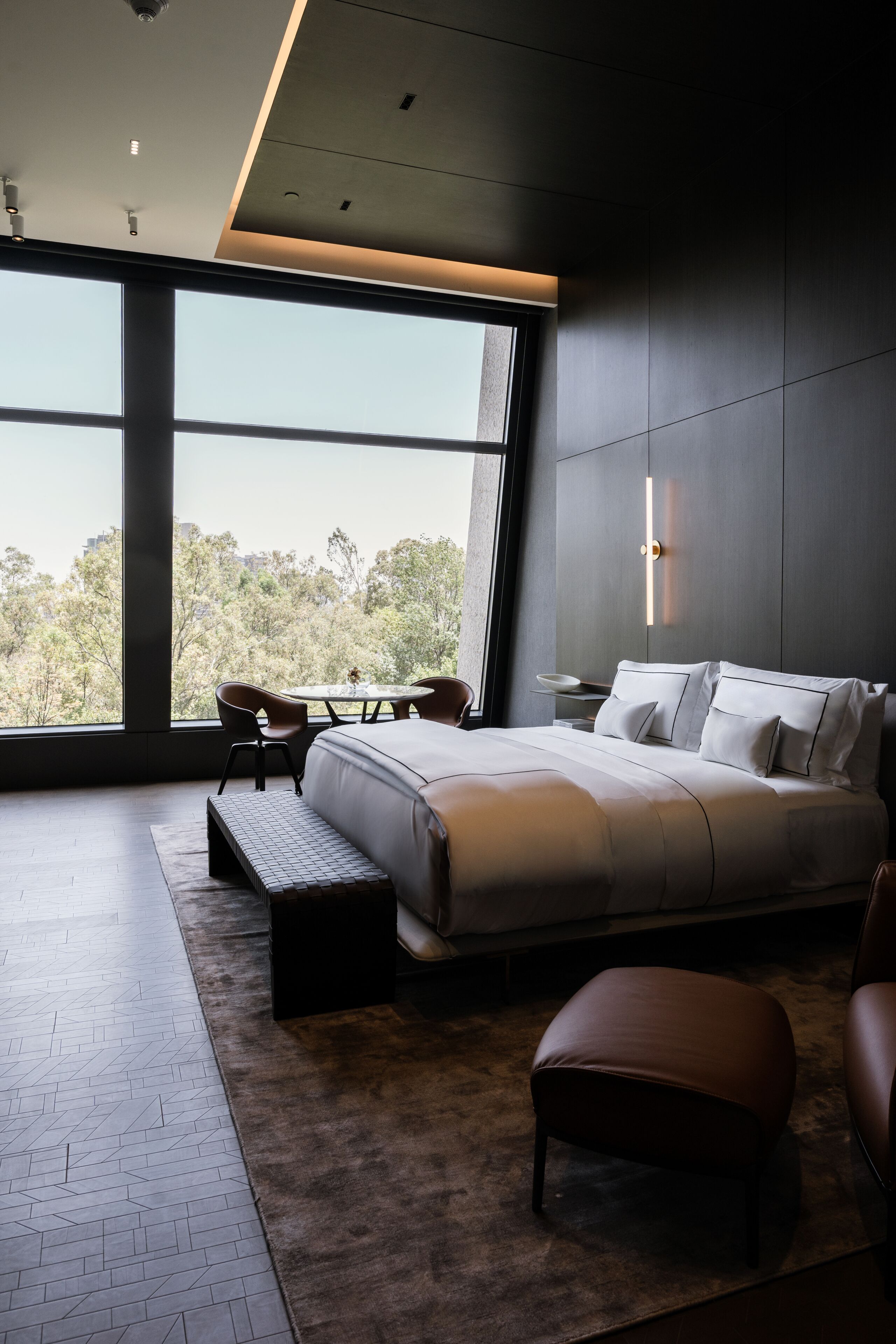 A modern bedroom with a large bed, a bench, and a view of trees through a large window.
