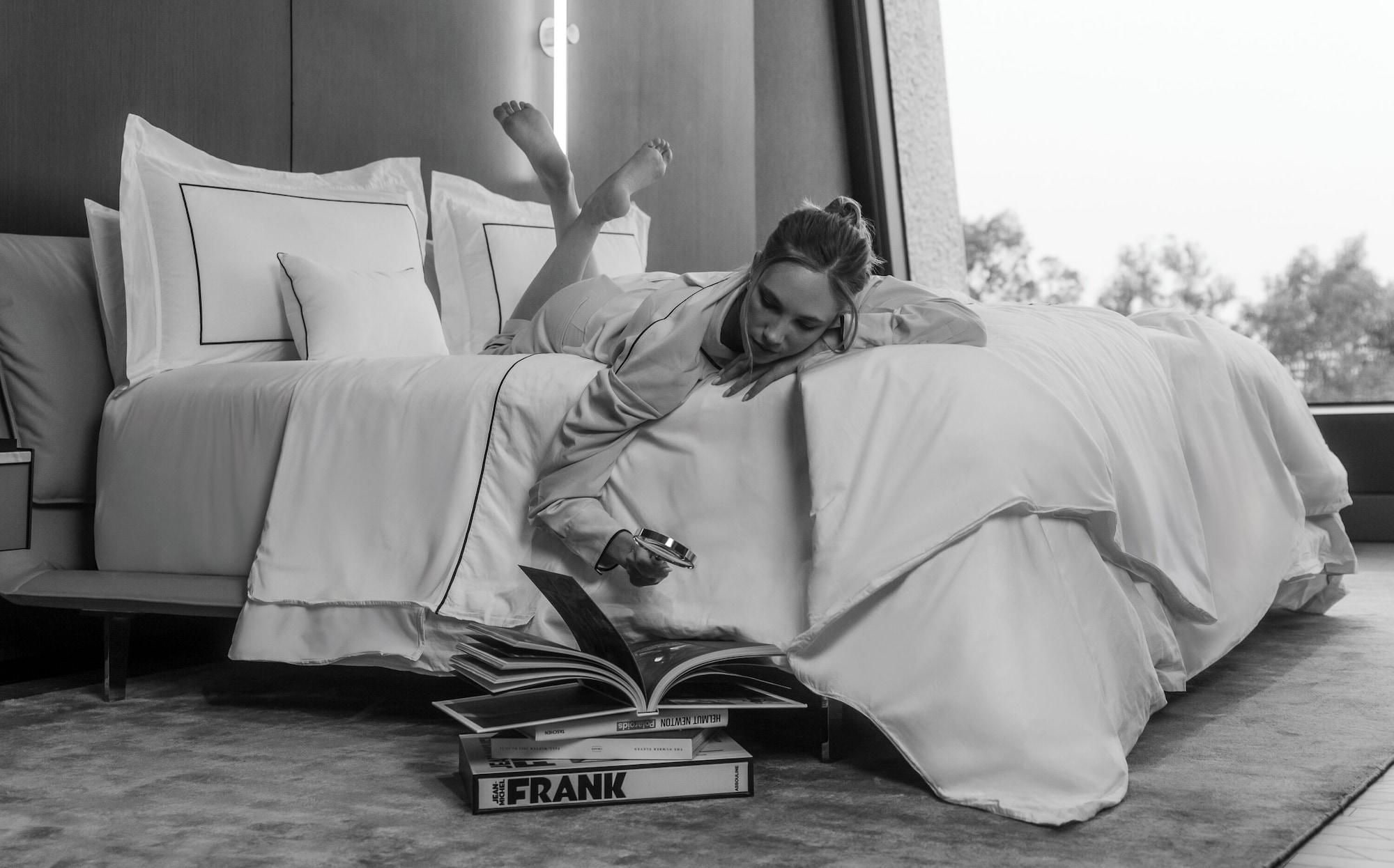 Una persona recostada en una cama en una habitación con un ventanal grande, leyendo un libro.