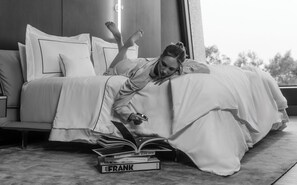 Eine Person liegt in einem Zimmer mit einem großen Fenster auf einem Bett und liest ein Buch.