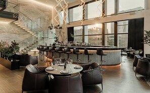A modern bar area with a high ceiling, a staircase, and a view of the city through large windows. - Hotel Alexander (Mexico City)