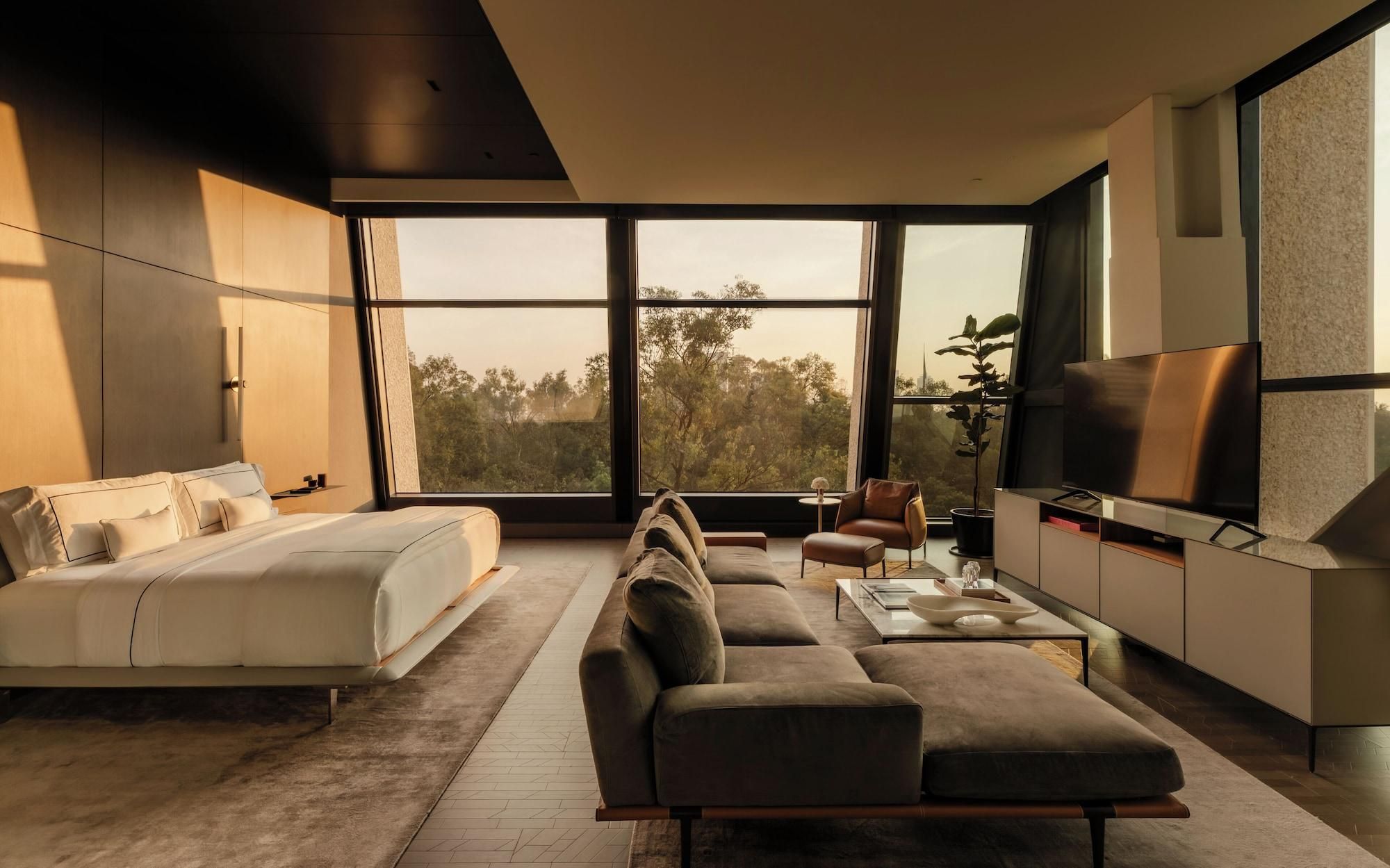 Une chambre moderne avec un grand lit, un canapé, une télévision et une vue sur les arbres à travers de grandes fenêtres.