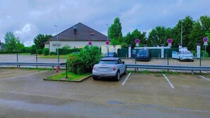 A parking area with multiple cars, a building with a sloped roof, and a green fence. - Premiere Classe Troyes Sud - Parc St Thibault (Bucheres)