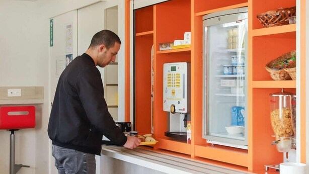 A man preparing food at a counter with a coffee machine and a refrigerator stocked with beverages. - Premiere Classe Troyes Sud - Parc St Thibault (Bucheres)