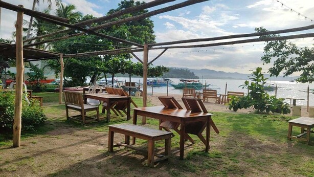 Un espacio al aire libre con mesas y sillas de madera, una estructura de pérgola y vistas al agua y a las embarcaciones.