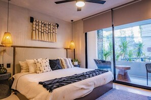 A modern hotel room with a large bed, two bedside lamps, a ceiling fan, and a view of the outdoors through large windows. - Bakal Tulum By Lockey (Tulum)