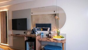 Una habitación de hotel moderna con una televisión de pantalla plana grande montada en la pared, un escritorio de madera con una cafetera, un refrigerador pequeño y un banco acolchado azul.