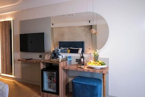 Una habitación de hotel moderna con una televisión de pantalla plana grande montada en la pared, un escritorio de madera con una cafetera, un refrigerador pequeño y un banco acolchado azul.