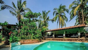 Una piscina envoltada de vegetació abundant i palmeres, amb una zona de seients coberta i un edifici al fons.